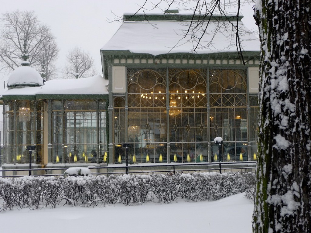 Glass building, snow on roof, chandelier glowing warmly inside.