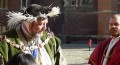 Large man in green velvet coat with gold chain around shoulders, black hat with white feathers trim, another man in red robe, no hat, in courtyard