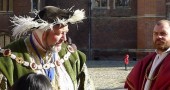 Large man in green velvet coat with gold chain around shoulders, black hat with white feathers trim, another man in red robe, no hat, in courtyard