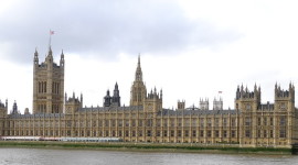 Long brown building with many pointed spires and a giant clock tower at the right hand end, on the far side of a broad river